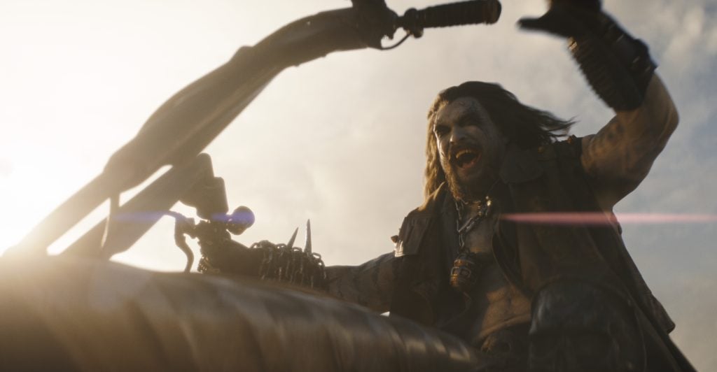 A wild-looking man with long hair rides a motorcycle, appearing to shout or yell, with his arm raised and sunlight illuminating the scene.