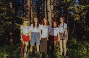 Five young women stand holding hands in a forest, all wearing matching "LEADER" t-shirts and shorts or pants, with trees and sunlight in the background.
