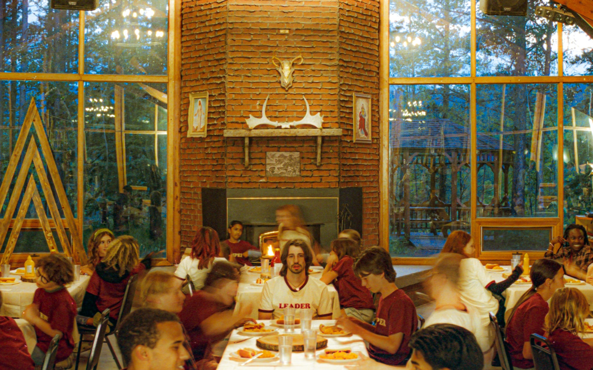 A group of people sit at long tables in a dining hall with large windows and a fireplace; one person in a "Leader" shirt sits at the center, facing the camera.