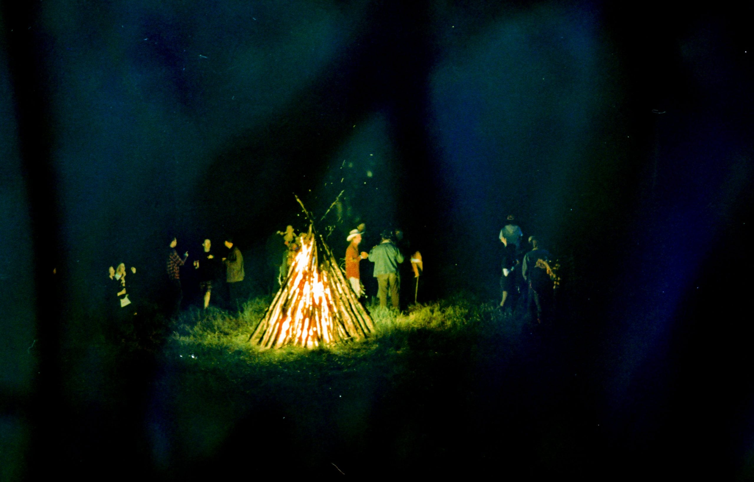 A group of people stand around a large, burning bonfire at night in an outdoor grassy area.