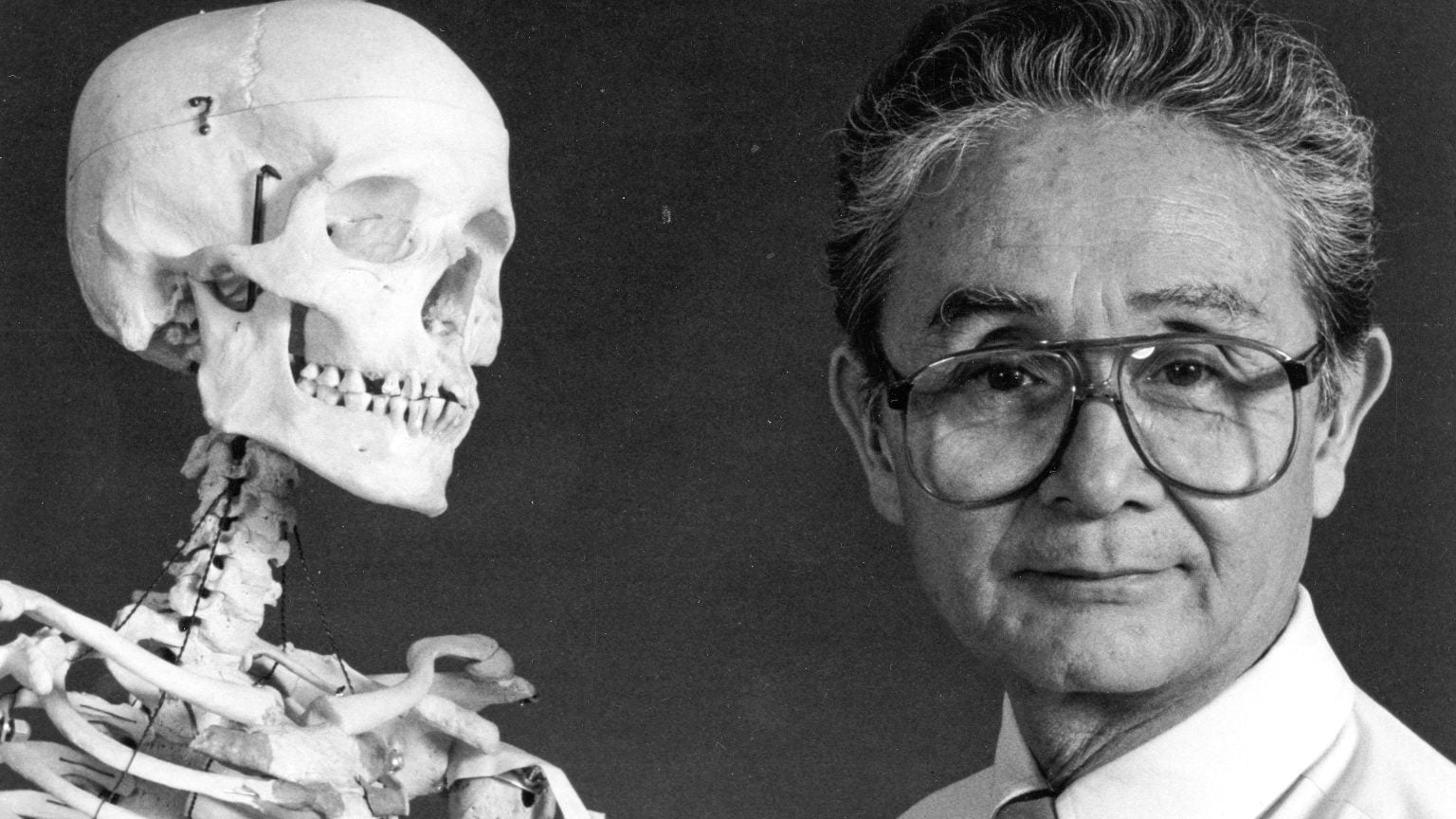 ‘Coroner To The Stars’ Review – Documentary Strips Away Sensationalism To Showcase Humanity An older man wearing glasses stands next to a human skeleton model, both facing the camera against a plain background.