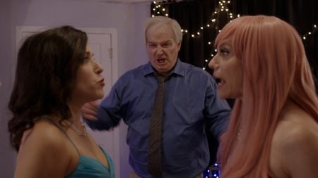 Season Two Of Short Form Comedy Series ‘Certifiable’ Debuts Trailer Ahead of Release (EXCLUSIVE) Two people in dresses argue face-to-face at a party while a man in a blue shirt and tie stands behind them, appearing angry. String lights hang in the background.