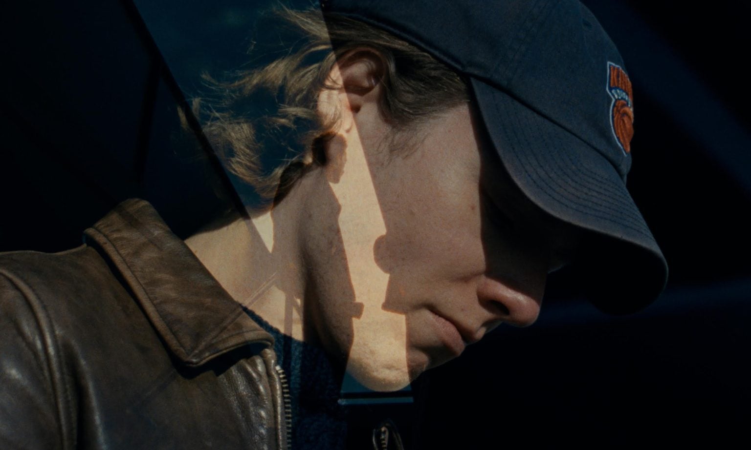 ‘Aqueduct’ Review – A Drama About More Than A Racetrack, But The People In The Crowd A person wearing a dark baseball cap and brown leather jacket is shown in partial shadow, with a double exposure effect featuring a silhouetted hand.