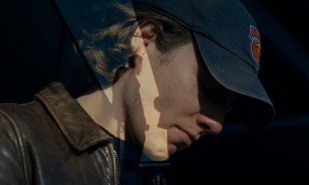 A person wearing a dark baseball cap and brown leather jacket is shown in partial shadow, with a double exposure effect featuring a silhouetted hand.
