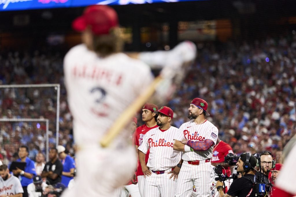 A baseball player swings in the foreground while three Philadelphia Phillies players and media personnel watch from the field during a game.