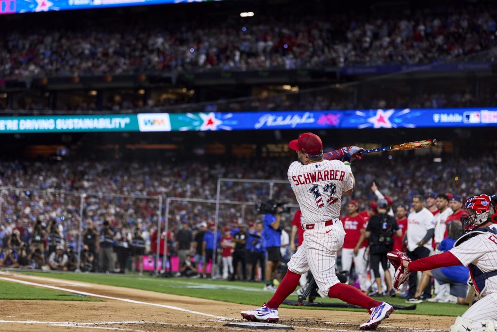 Baseball player in a pinstriped uniform swings a bat during a game, with a large crowd in the background and another player crouched behind the plate.
