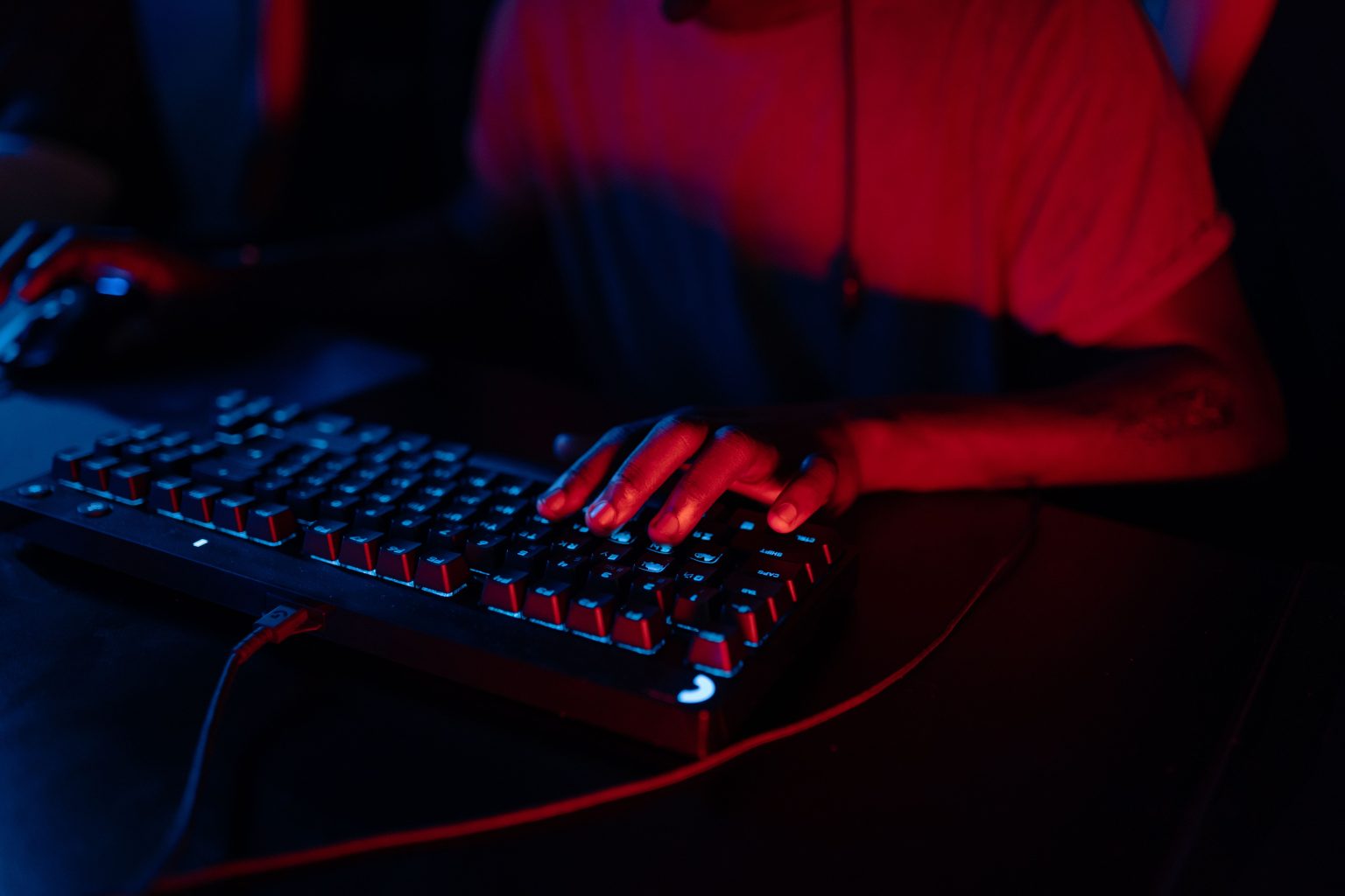 Person typing on an illuminated mechanical keyboard in a dimly lit room, with red and blue lighting highlighting their hands and shirt.