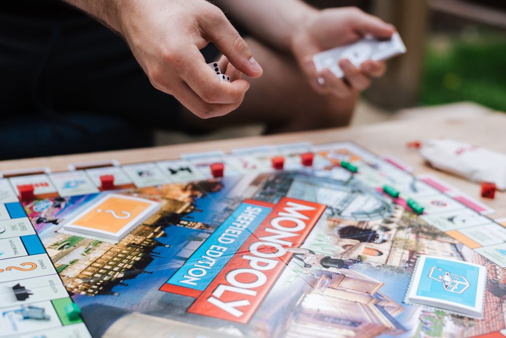 Person rolling dice and holding cards while playing the Monopoly Sheffield Edition board game, with game pieces and cards arranged on the board.