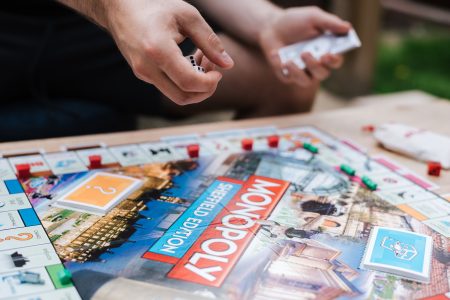 Legacy Entertainment Intellectual Properties Anchor Latest Wave Of Online Slot Releases Person rolling dice and holding cards while playing the Monopoly Sheffield Edition board game, with game pieces and cards arranged on the board.