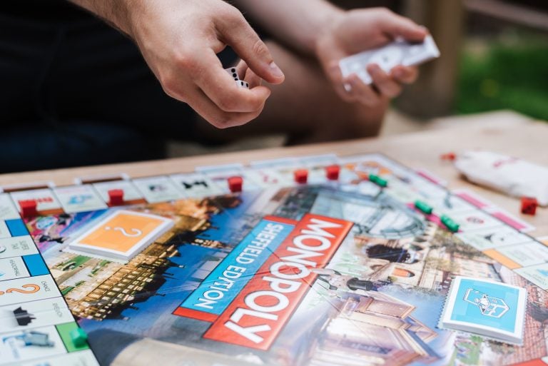 Legacy Entertainment Intellectual Properties Anchor Latest Wave Of Online Slot Releases Person rolling dice and holding cards while playing the Monopoly Sheffield Edition board game, with game pieces and cards arranged on the board.