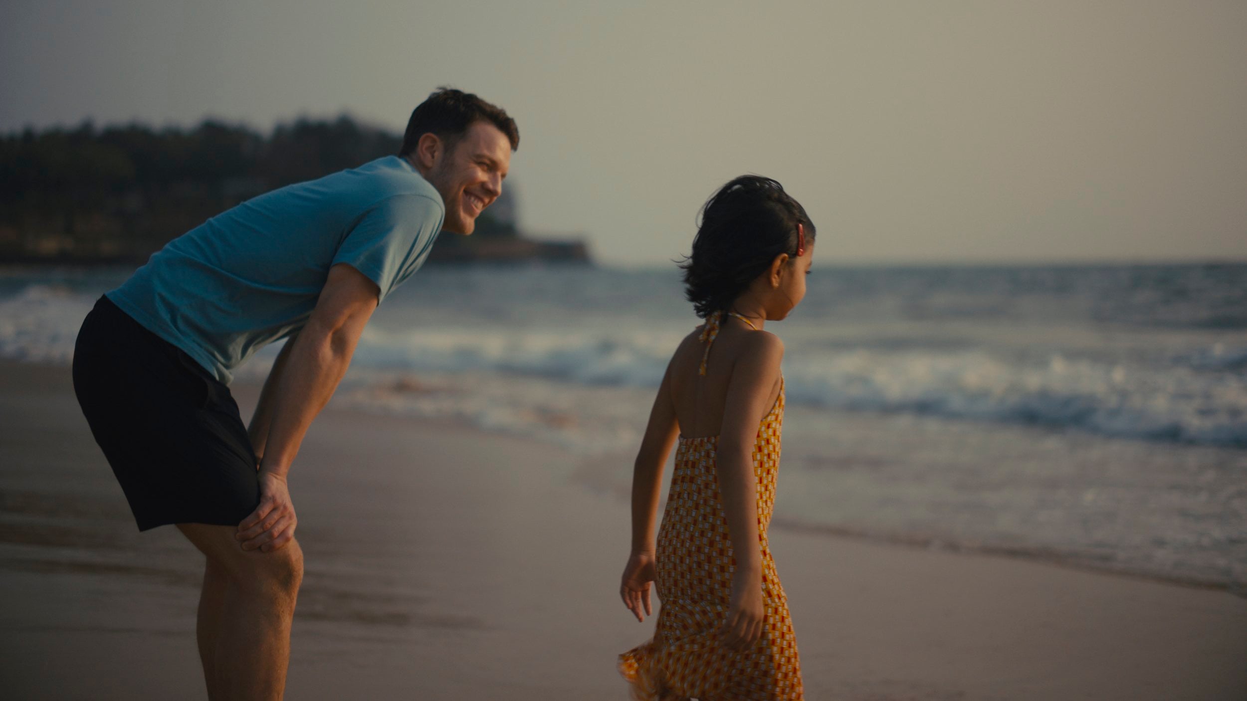An adult and a child stand near the shoreline at the beach, looking toward the ocean under soft, evening light.