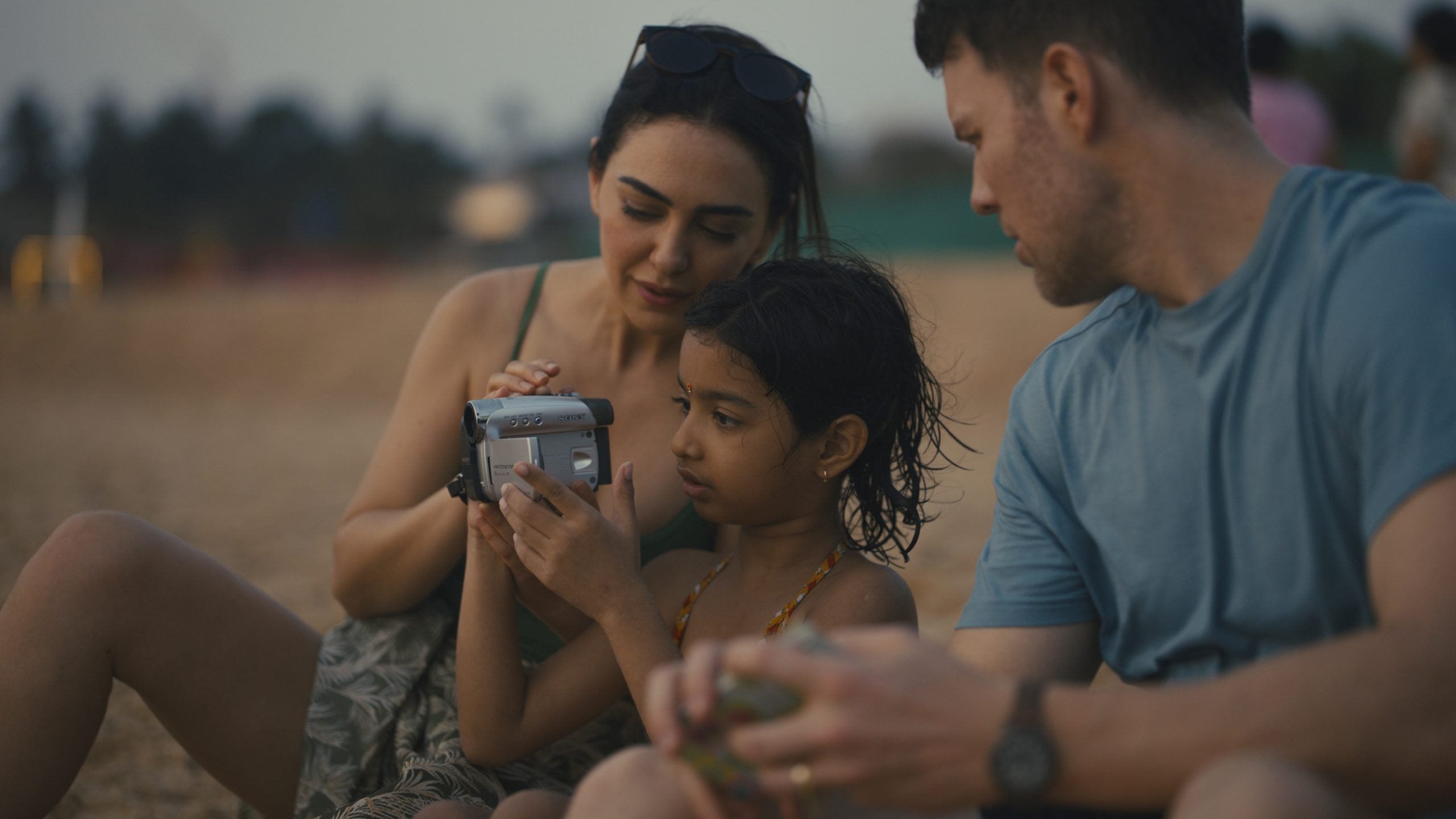 A woman and girl look at a handheld camera together on a beach while a man sits beside them holding an object, all appearing relaxed.