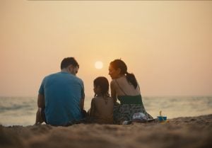 Three people sit on a sandy beach facing the ocean at sunset, with a toy bucket and shovel nearby.
