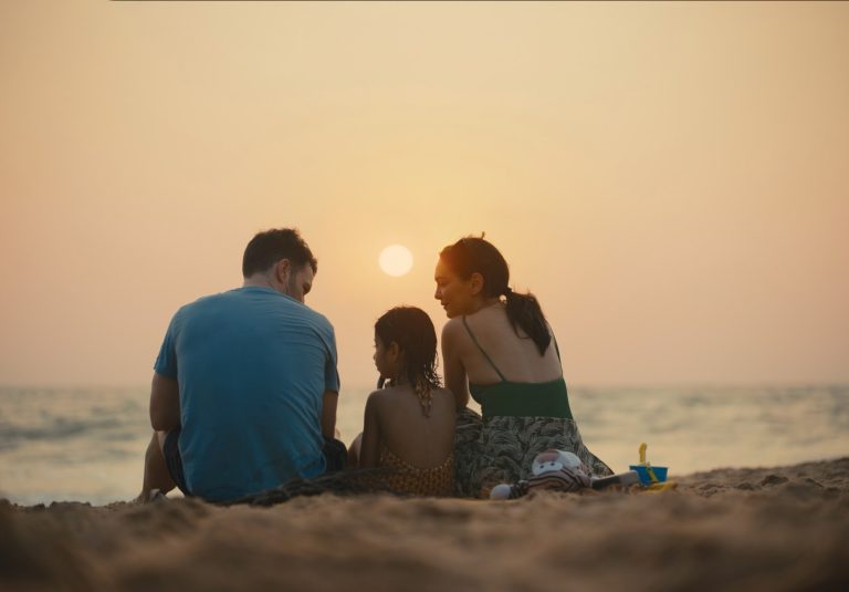 Three people sit on a sandy beach facing the ocean at sunset, with a toy bucket and shovel nearby.