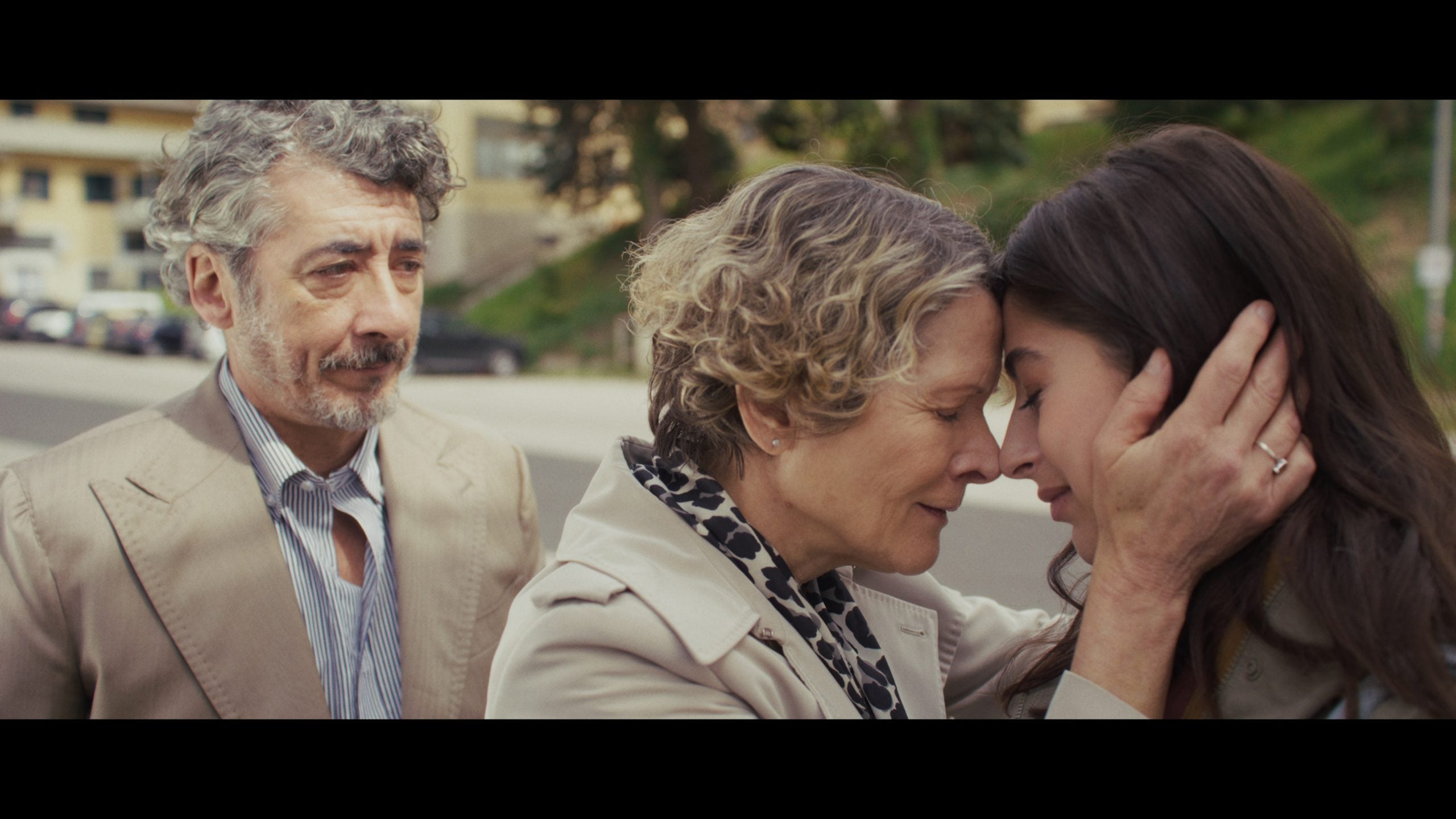 A woman gently holds a younger woman's face, their foreheads touching, while a man stands nearby watching them. All three are outdoors, dressed in light-colored clothing.
