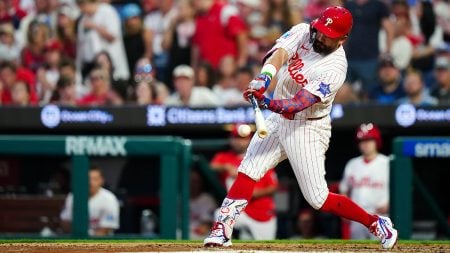 Netflix’s Home Run Derby: Who Is In? Who Will Win? Home Run Derby - A Philadelphia Phillies player in a white pinstriped uniform swings a bat at a baseball during a game, with spectators in the background.