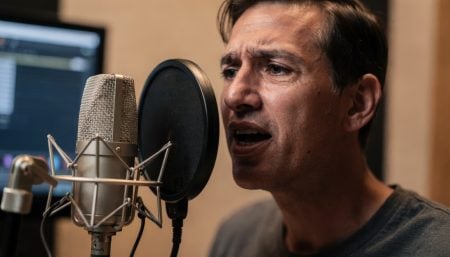 Beyond The Booth: How Modern ADR, Voiceover Integration, And Remote Tracking Are Reshaping Global Post-Production A man speaks or sings into a microphone in a recording studio, positioned close to a pop filter, with a computer screen visible in the background.