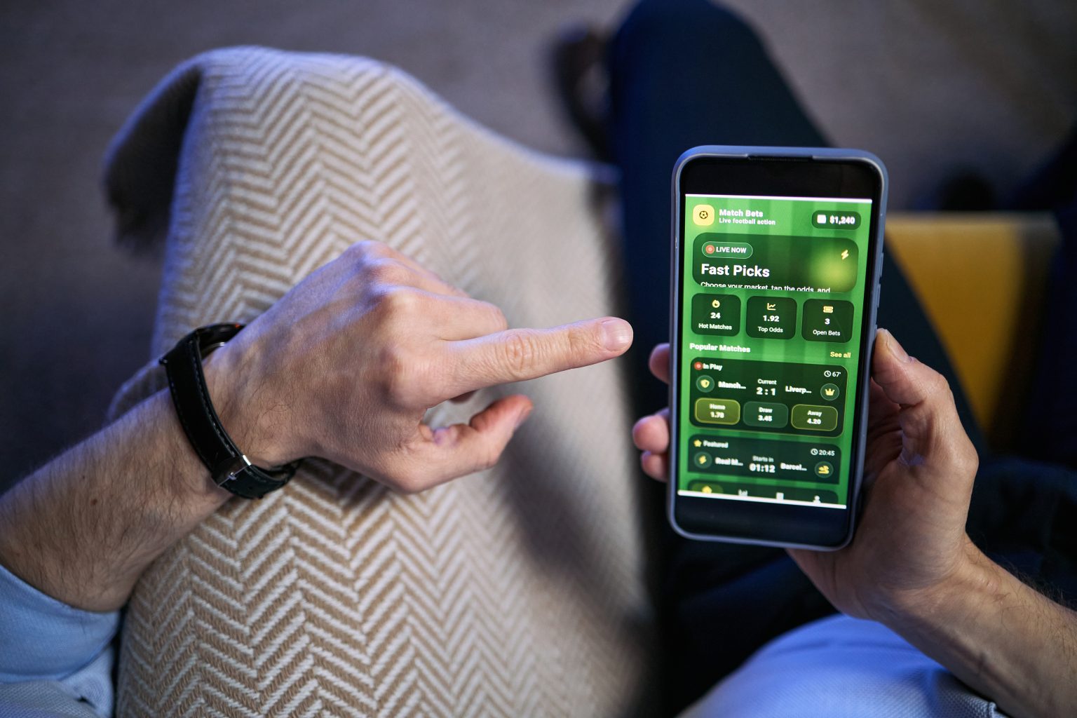 How An Online Sportsbook Site Adds To The Gaming Experience A person sitting on a couch uses a smartphone displaying a sports betting or gambling app with various betting options visible on the screen.
