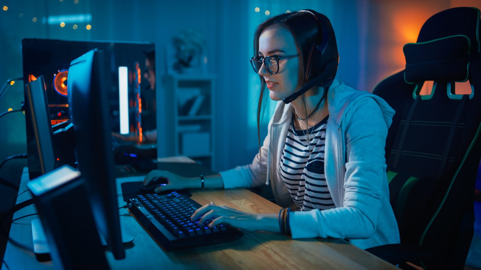 A person wearing headphones and glasses sits at a desk, using a computer and playing a game in a dimly lit room.