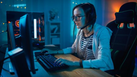 The Hidden Causes of Gamer Eye Strain A person wearing headphones and glasses sits at a desk, using a computer and playing a game in a dimly lit room.
