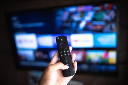 Why My IPTV Kept Buffering On A Fast Connection — And How I Finally Fixed It on A Firestick A hand holds a remote control pointed at a television screen displaying a streaming service menu with various show options.