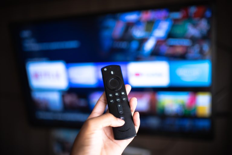 A hand holds a remote control pointed at a television screen displaying a streaming service menu with various show options.