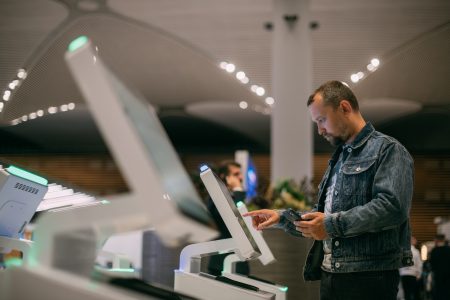 A man uses a self-service kiosk at an airport, holding his phone and interacting with the touchscreen.