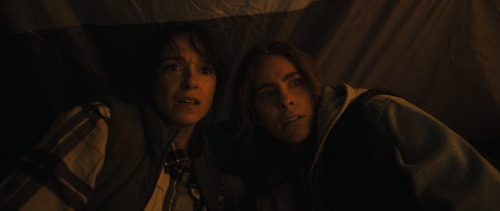Two people sit close together in dim lighting, looking ahead with concerned expressions; a tent-like fabric is visible in the background.