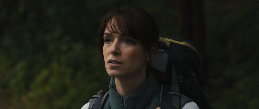 A woman with brown hair and bangs, wearing a jacket and backpack, stands outdoors in front of blurred greenery.