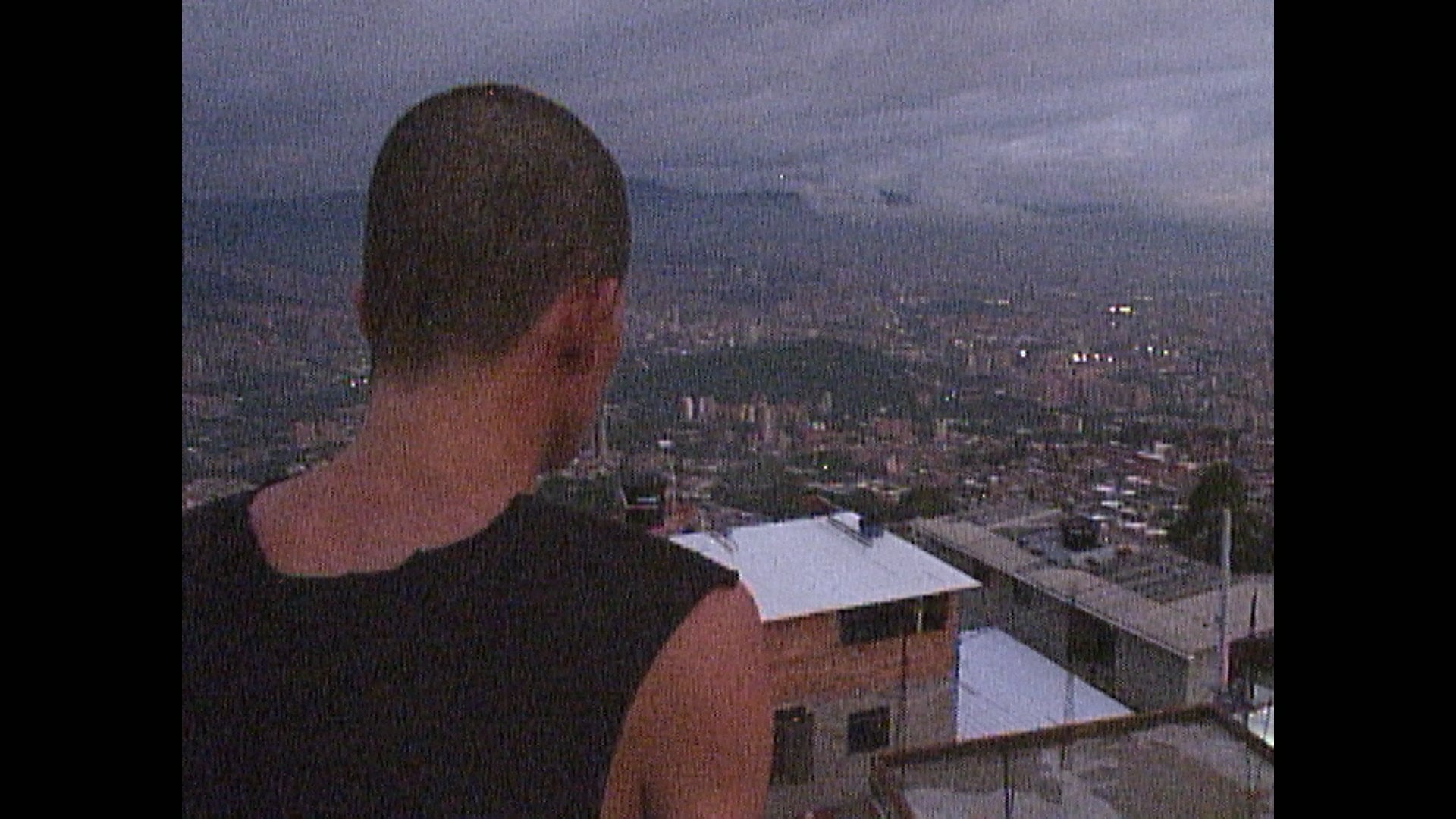 A person with close-cropped hair and a dark sleeveless shirt stands on a rooftop, looking out over a cityscape at dusk.