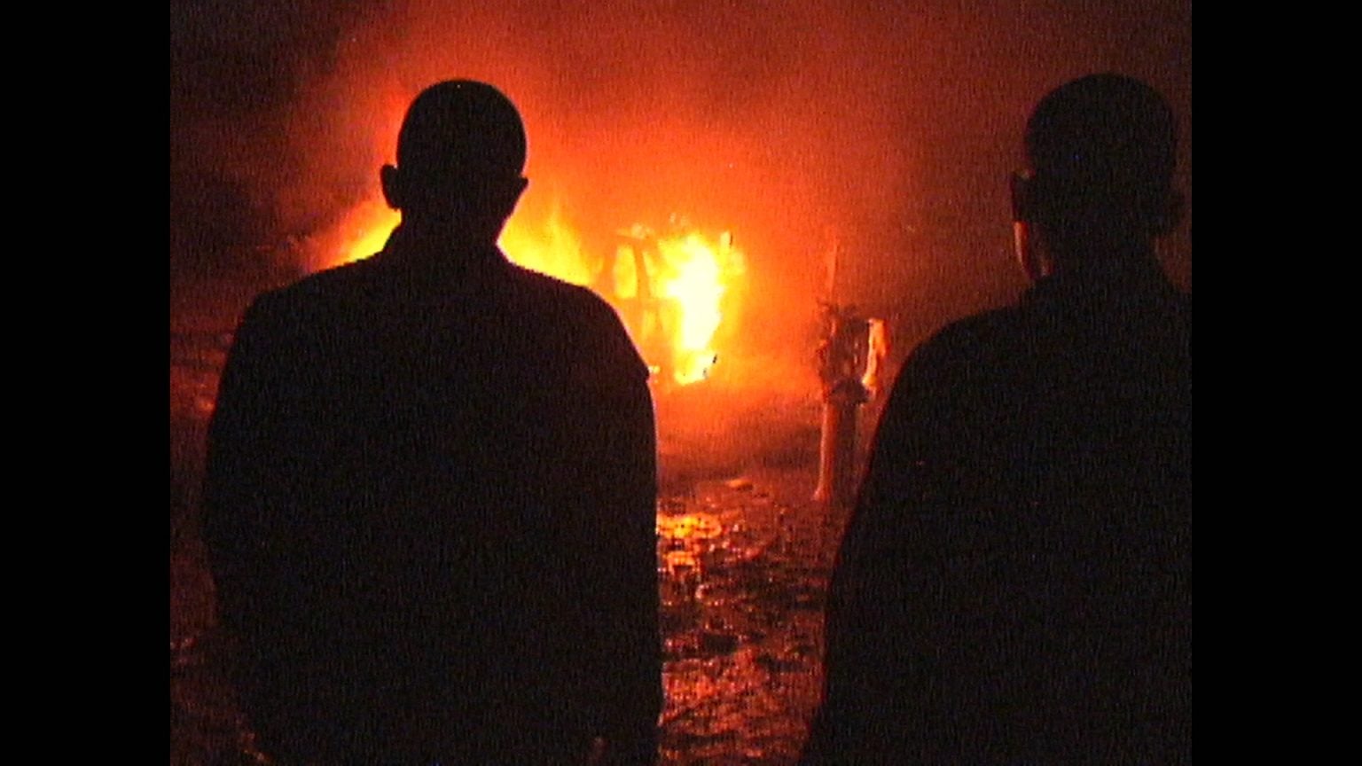 Two silhouetted figures stand facing a large fire at night, with flames and smoke illuminating the scene in the background.