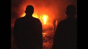 ‘Barrio Triste’ Review – A Found Footage Film That Takes Bold Swings & Evokes Pathos Two silhouetted figures stand facing a large fire at night, with flames and smoke illuminating the scene in the background.