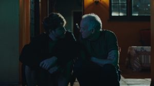 Two men sit on a porch at night, facing each other and having a serious conversation. Warm indoor light contrasts with the dark outdoor setting.