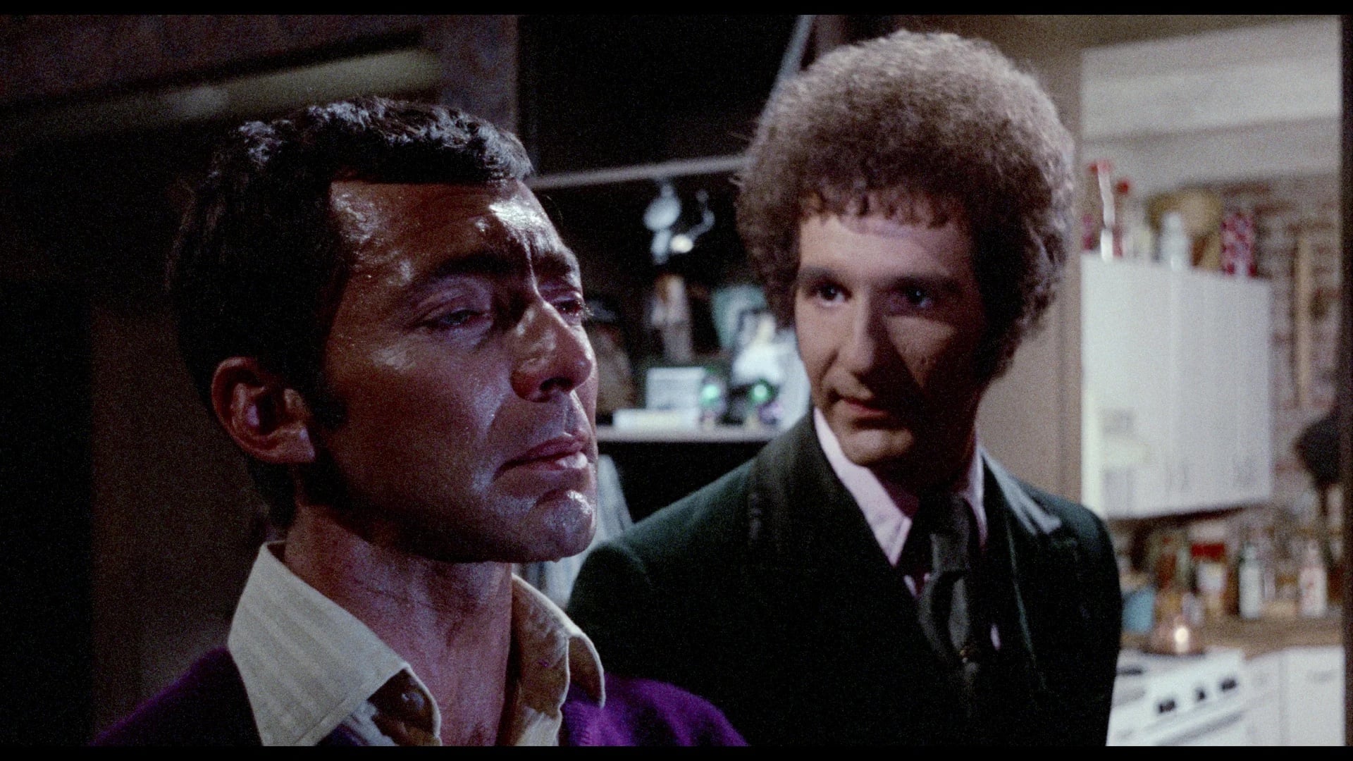 Two men stand indoors; one looks emotional and faces left, while the other, wearing a suit, looks at him from behind. The background shows a cluttered kitchen.
