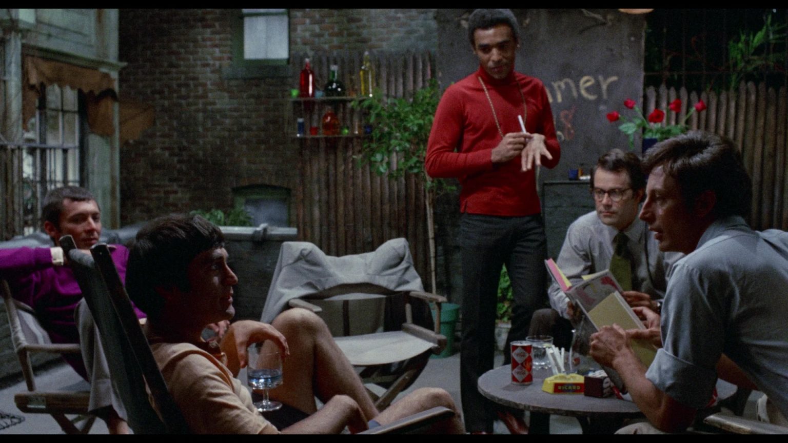 ‘The Boys In The Band’ (1970) 4K UHD Blu-Ray Review – An Unexpected & Engrossing Early Effort From William Friedkin Five men sit and stand around a table in a dimly lit outdoor patio, talking. Some hold drinks, and books are visible on the table. The background features a fence and wall with writing.