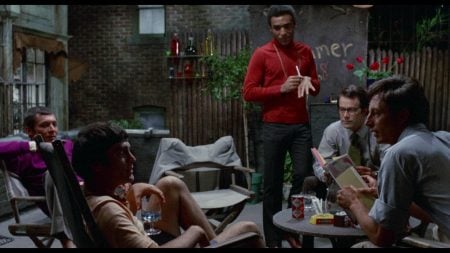 Five men sit and stand around a table in a dimly lit outdoor patio, talking. Some hold drinks, and books are visible on the table. The background features a fence and wall with writing.