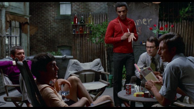 ‘The Boys In The Band’ (1970) 4K UHD Blu-Ray Review – An Unexpected & Engrossing Early Effort From William Friedkin Five men sit and stand around a table in a dimly lit outdoor patio, talking. Some hold drinks, and books are visible on the table. The background features a fence and wall with writing.