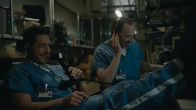 Two people in blue scrubs sit in a dimly lit room, wearing headsets and talking on phones, surrounded by electronic equipment and wires.