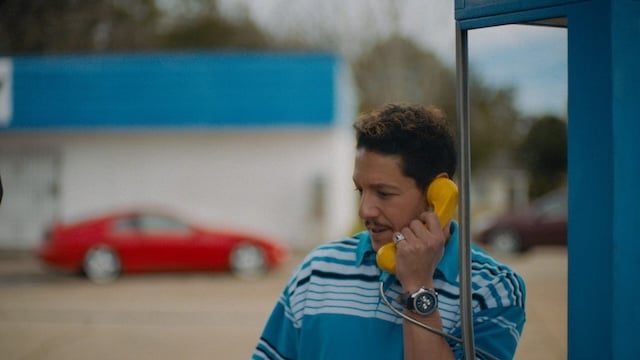 A man in a blue striped shirt uses a yellow payphone outside, with a red car and a blue-and-white building in the background.