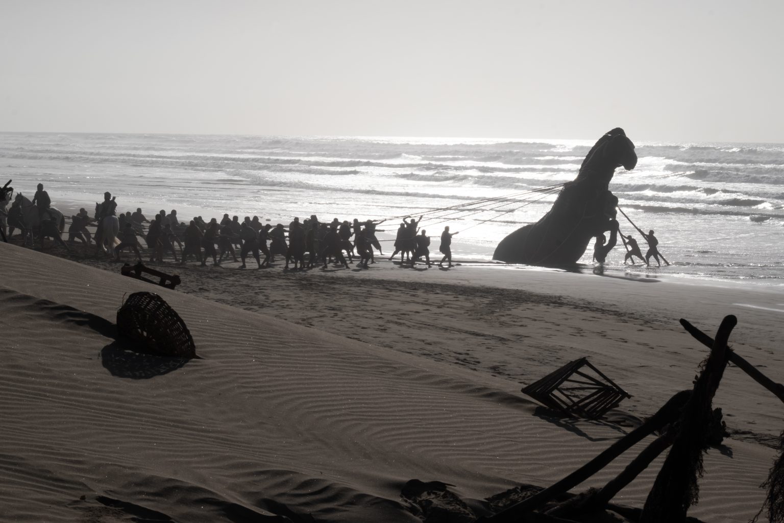 A group of people on a beach pull ropes attached to a large dinosaur statue near the shoreline, with scattered debris in the sand.
