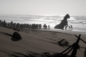 Christopher Nolan’s Biggest Box Office Hit Is No Longer A Batman Movie A group of people on a beach pull ropes attached to a large dinosaur statue near the shoreline, with scattered debris in the sand.