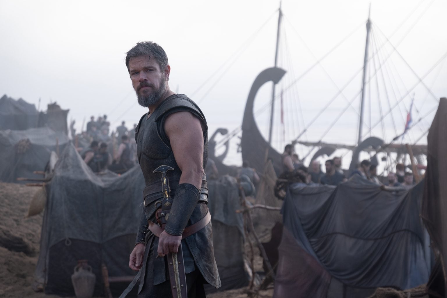 ‘The Odyssey’ Review – A Breathtaking Epic Of Hubris and Humility A man in medieval armor stands with a sword on a battlefield near tents and Viking-style ships, with people gathered behind him under a cloudy sky.