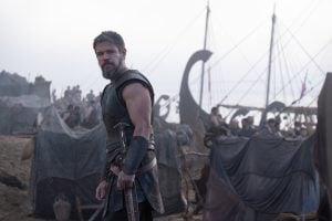 ‘The Odyssey’ Review – A Breathtaking Epic Of Hubris and Humility A man in medieval armor stands with a sword on a battlefield near tents and Viking-style ships, with people gathered behind him under a cloudy sky.
