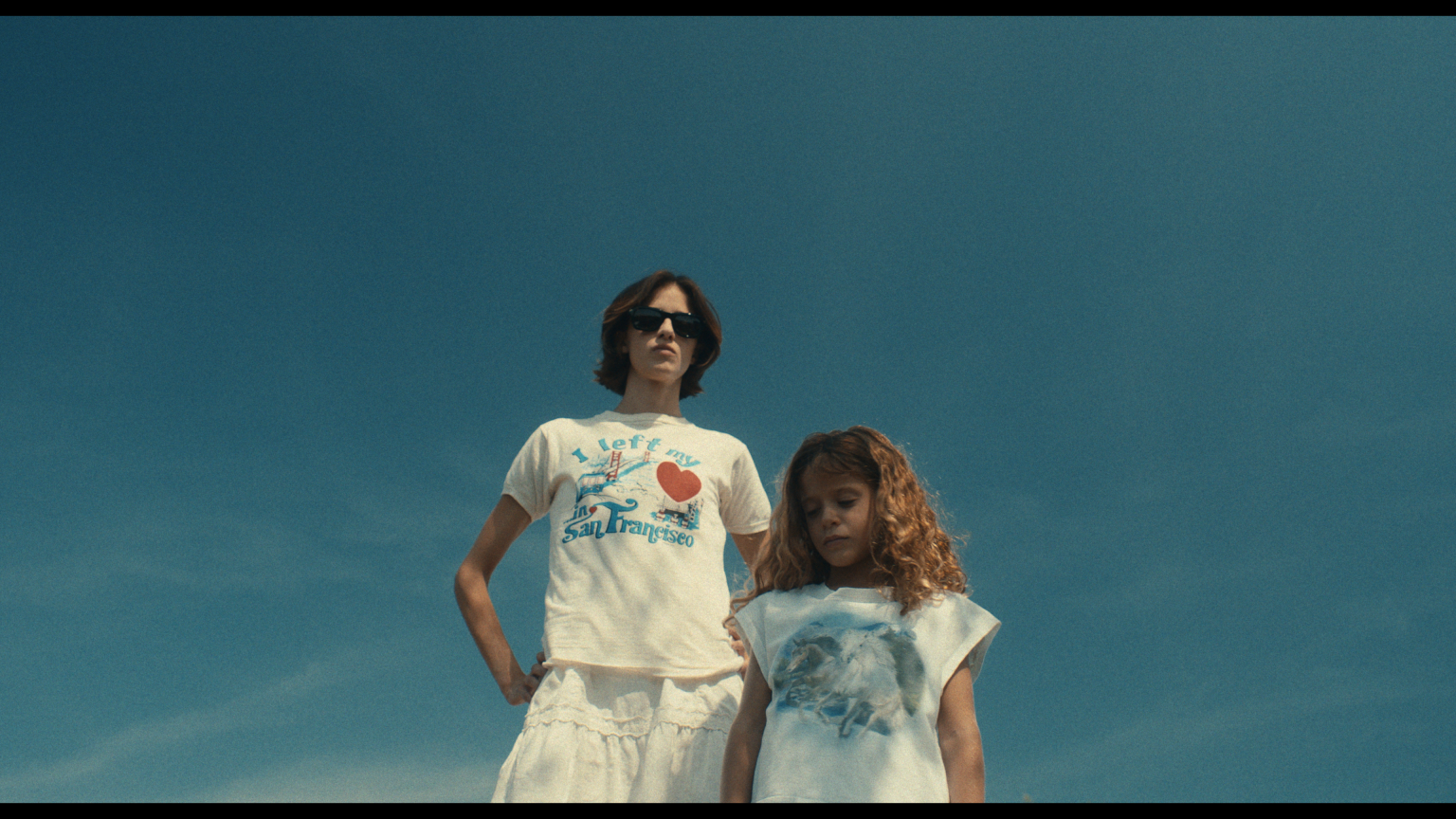 Two children stand outdoors against a clear blue sky. The older child wears sunglasses and a white "I Left My Heart in San Francisco" shirt; the younger child wears a white shirt with a graphic.