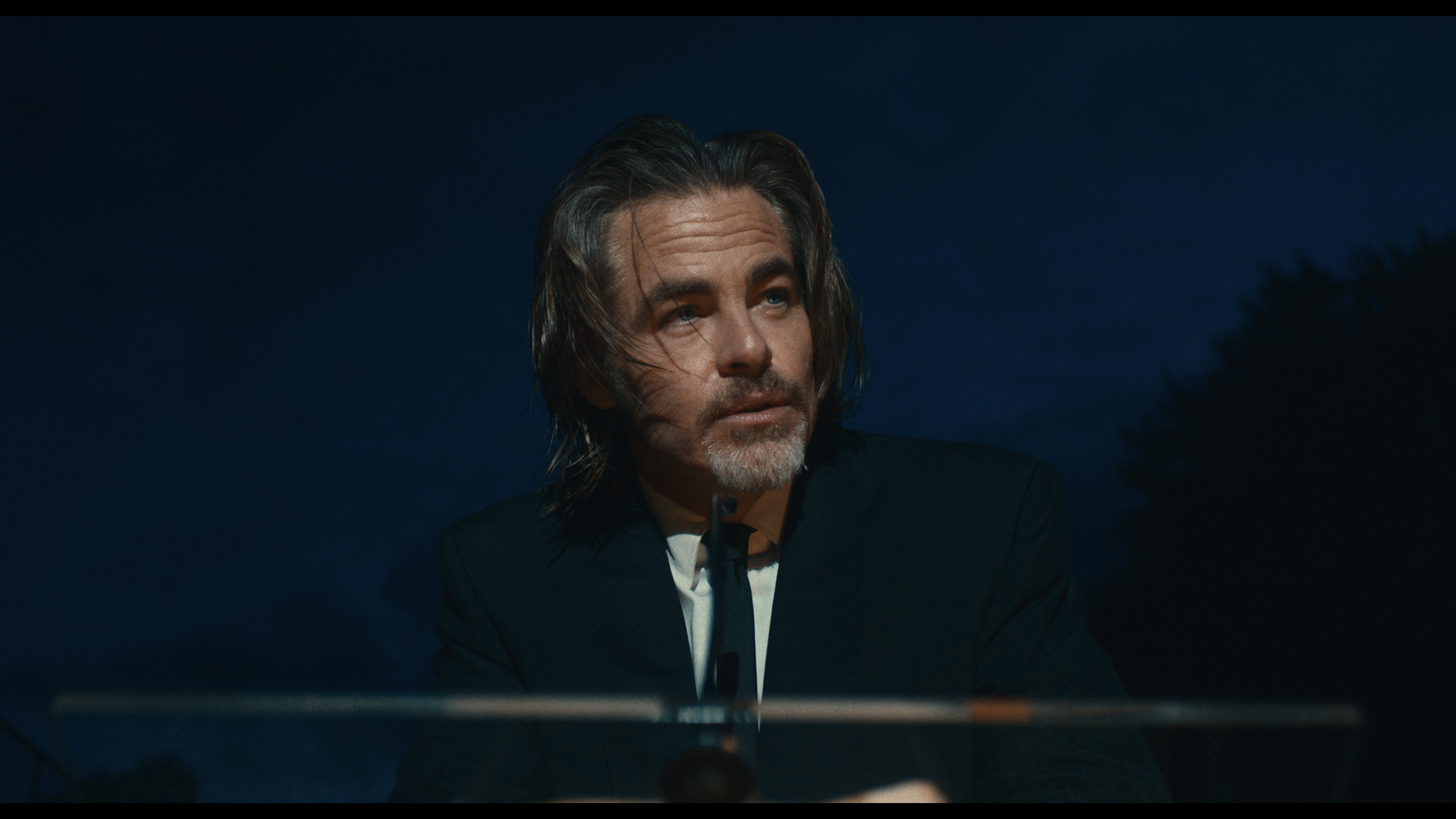 A man with shoulder-length hair and a beard, dressed in a dark suit, sits behind a lectern outdoors at dusk, looking slightly to the side.