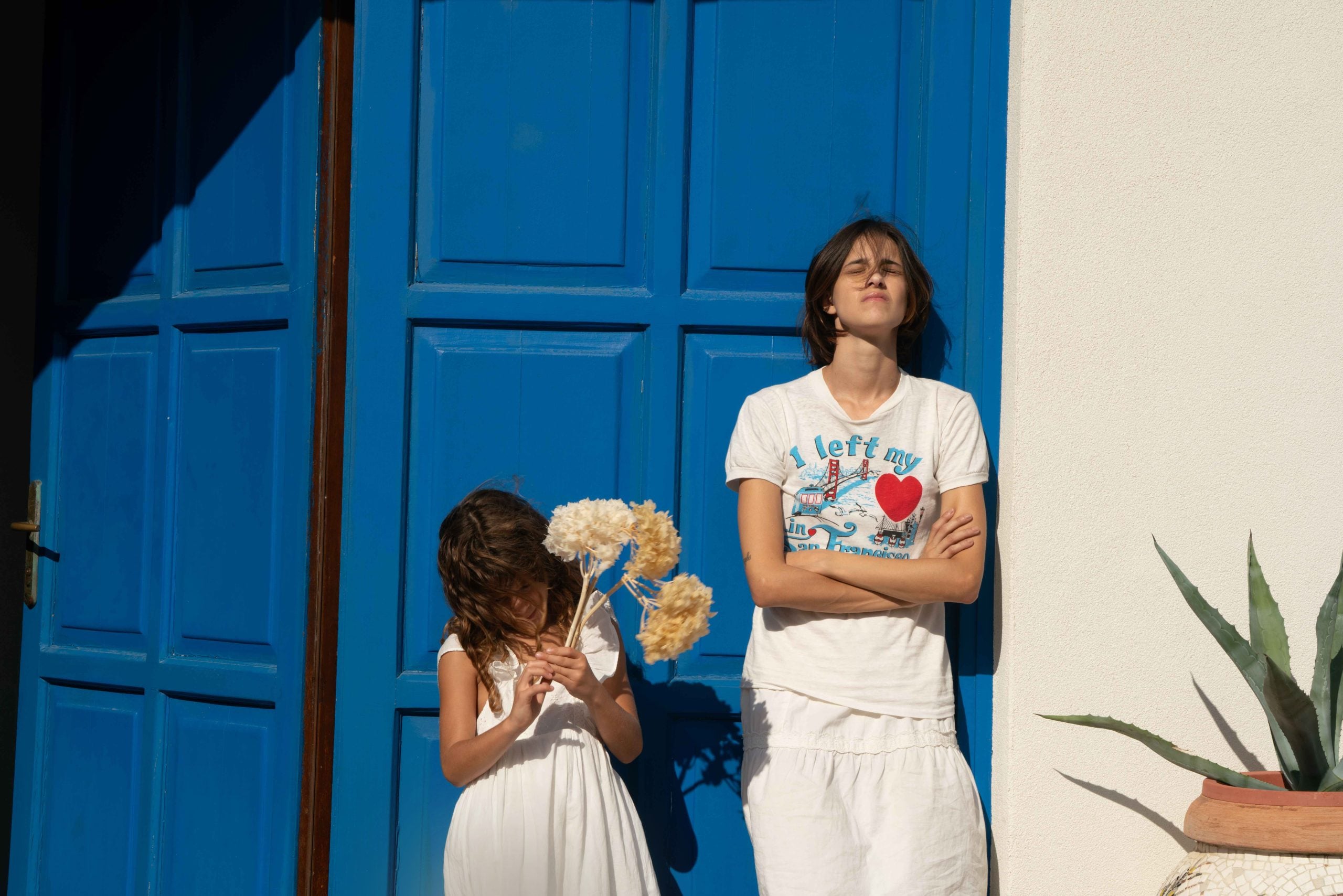 Two people in white clothes stand in sunlight by a bright blue door; the taller person has arms crossed and eyes closed, the shorter holds beige flowers and looks down.