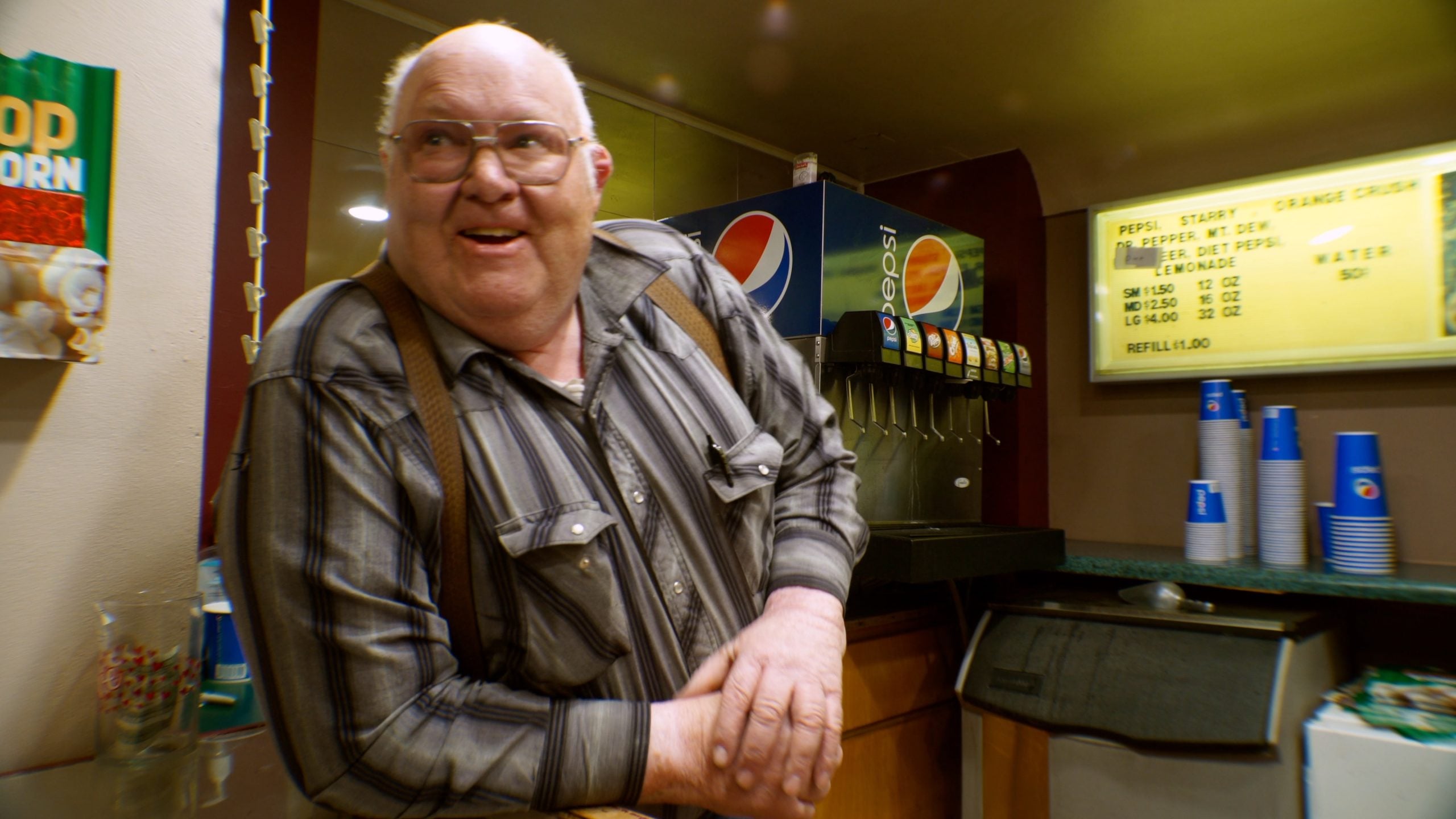 A man in a striped shirt leans on a counter near a soda fountain and a popcorn sign in what appears to be a concession stand area.
