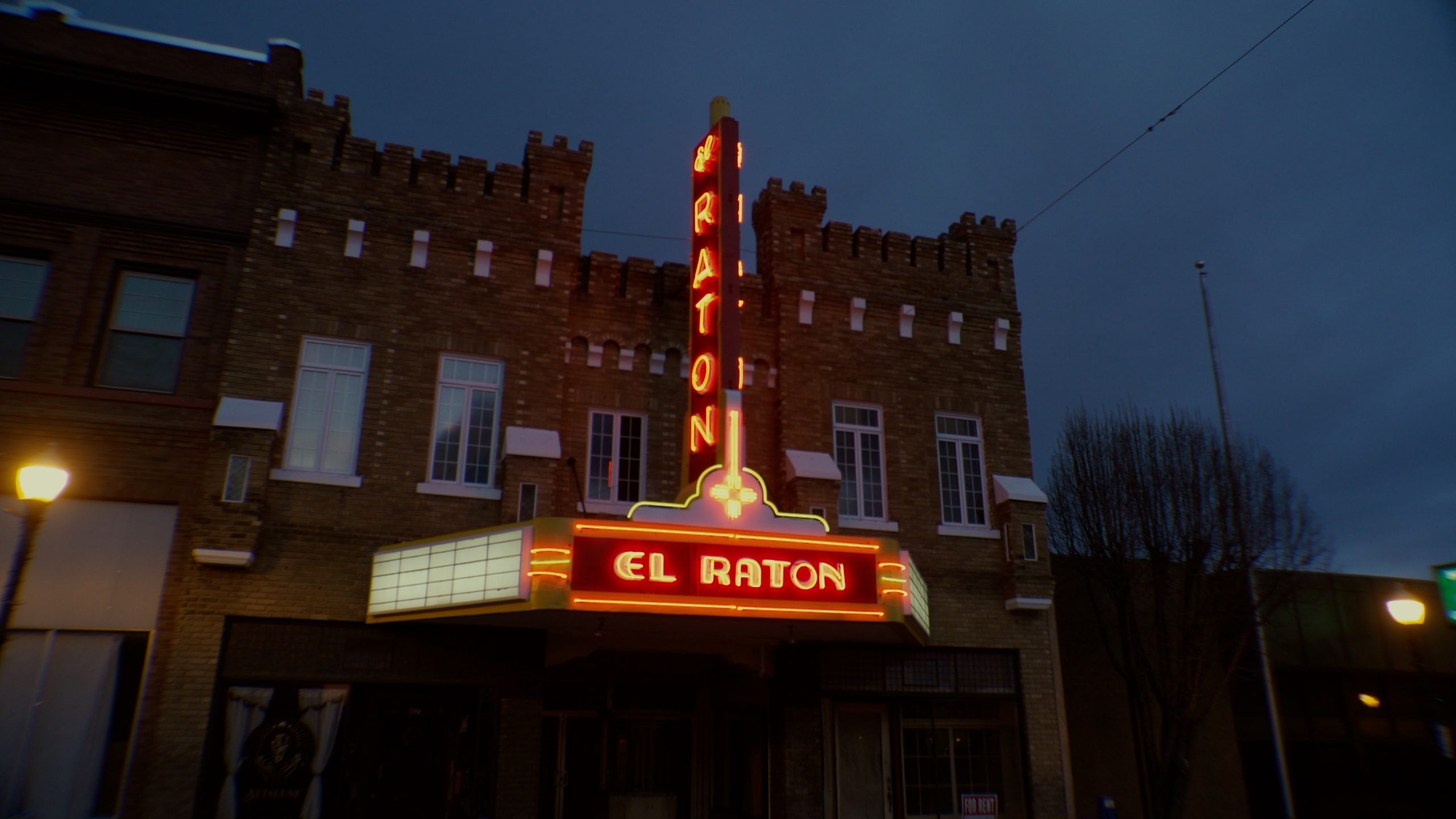 The El Raton theater building at night with its neon sign lit in red and yellow, displaying "El Raton" vertically and horizontally on the marquee.
