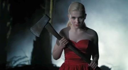 A woman in a red dress stands outdoors at night, holding an axe with both hands and looking forward, with mist and trees in the background.