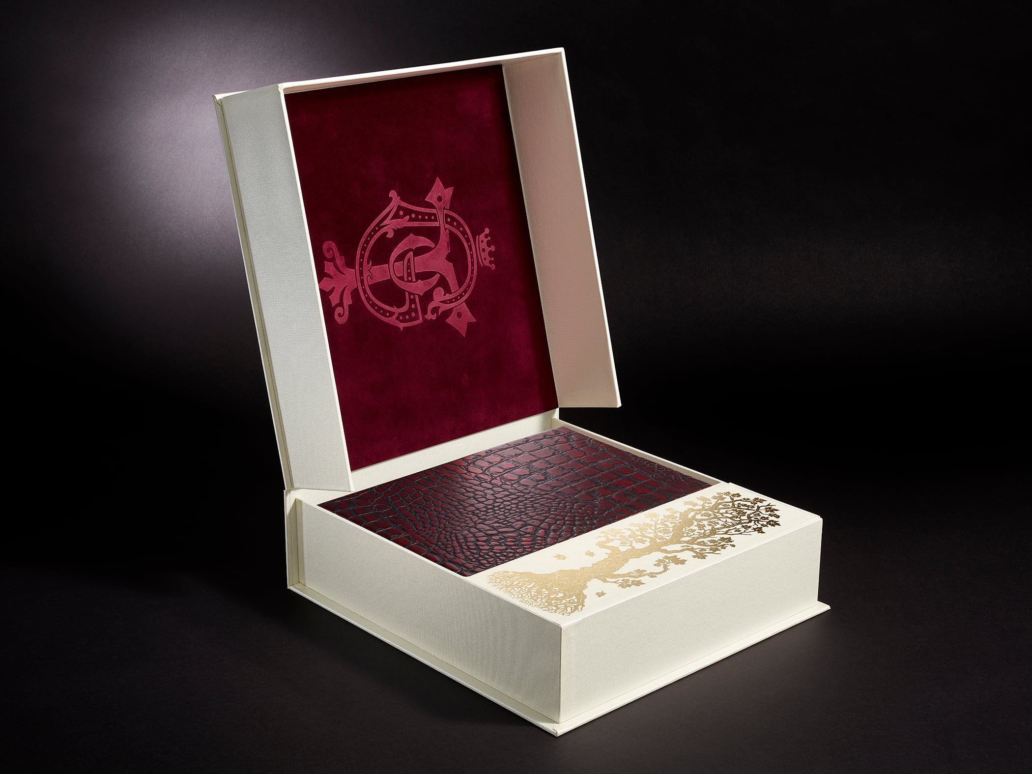 A decorative book with a red textured cover and gold design is displayed in an open cream-colored box with a red velvet interior and embossed emblem.